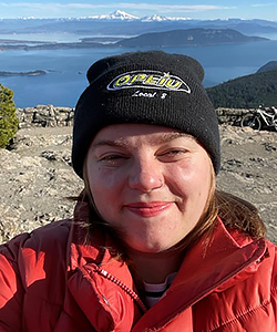Selfie of a young white woman wearing a black OPEIU beanie and a red winter jacket, with the view of a glaciated mountain and a blue body of water in the distance behind her