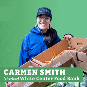 Smiling woman wearing a ball cap, blue jacket, and glasses, holding boxed of produce with the label CARMEN SMITH (she/her) White Center Food Bank