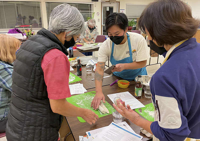 Three Asian women in aprons and masks prep food around a table in a training.
