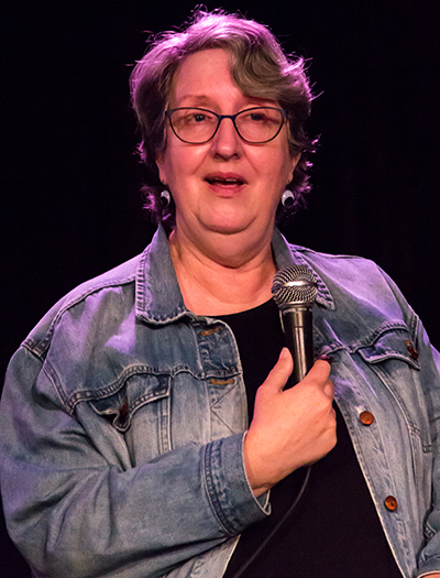 Wearing a denim jacket over a black dress, Kim McGillvray speaks into a microphone.