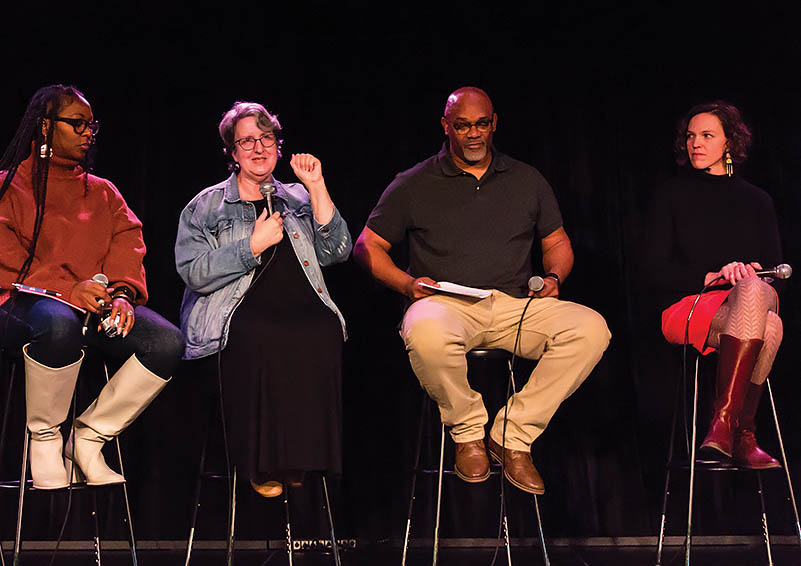 Four people sit on stools on a stage. Kim McGillivray, wearing a jeans jacket over a black dress, is speaking while the three other people listen.
