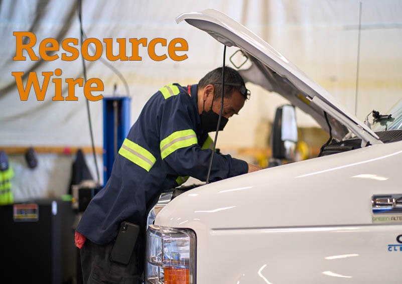 A mechanic in a blue shirt with reflective yellow stripes looks under the hood of a white vehicle. The words "Resource Wire" appear in orange above him.