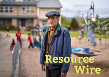 A man in a blue coat and cap stand in front of a playground.