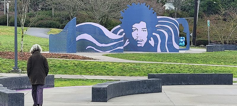 A purple-hued sculpture depicting the face of Jimmi Hendrix with undulating wages of purple and white emanating outward. It sits in a park with green grass and black stone benches.