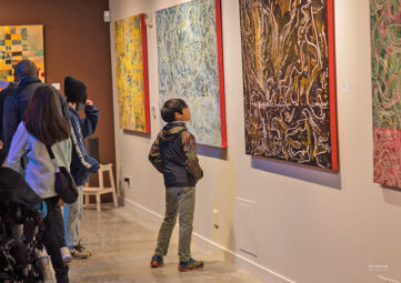 A small boy gazes at one of several colorful abstract paintings on the walls of a gallery room. A small crowd of people can be seen off to the left of the image.