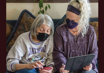 Elderly woman holds a cellphone as a younger senior woman with a tablet looks on. Both wear masks and have grey hair.