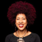 Portrait of a Black woman with a reddish afro, wearing a black top and wood and metal necklace.