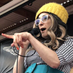 Photo of a white woman with glasses speaking into a microphone and pointing her forefinger. She wears a bright yellow hat, wearing a black and white checkered dress with a big aqua belt.