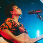 Portrait of a Black woman playing guitar and singing into a microphone, wearing an orange and black top.