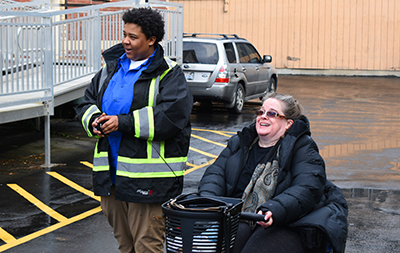 A woman in a reflective jacket and a woman in a motorized wheelchair laugh as they watch something off camera. 