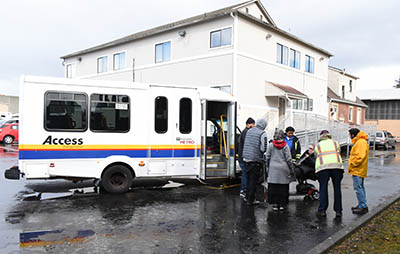 A group of seven people gather in a huddle just outside a paratransit van in front of a white building.