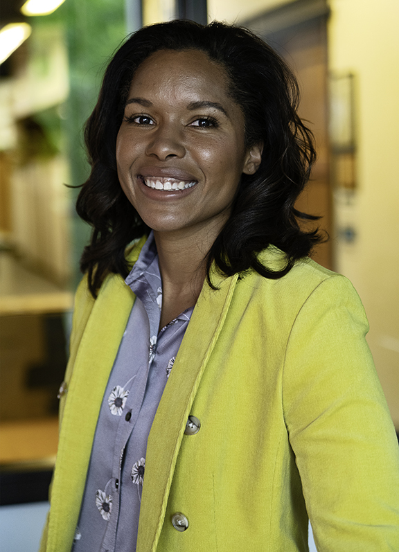 Portrait of a smiling woman with black hair wearing a sage-green blouse and a bright yellow blazer.