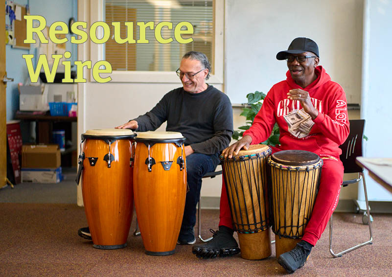 Two men smiling and playing drums with their hands in an office setting. The words "Resource Wire" appears besides them.