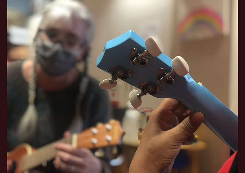 Foreground: A child's hand holds a chord on the neck of a bright blue ukulele. Blurred background: A grey-haired woman with glasses, braids, and a grey mask on holds a chord on a wooden ukulele.