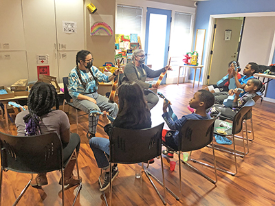 Children holding ukuleles are seated in a semi-circle around two adults also holding ukuleles.