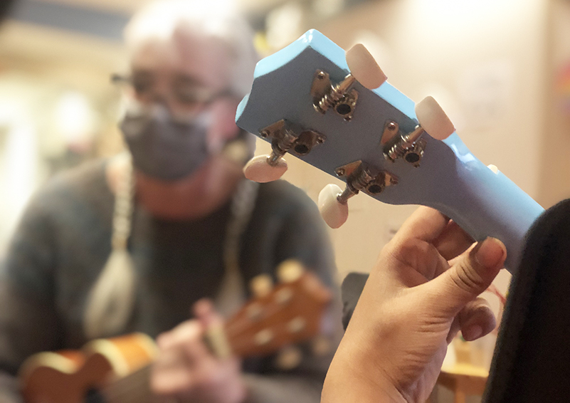 Image showing a child's hands fingering the frets of a power-blue ukulele. in the background, out of focus, a woman with long white hair is holding another ukulel.