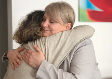 Woman with short blonde hair hugs a woman with shoulder-length curly brown hair. The corner of a colorful painting shows in the upper right.