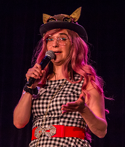 A woman holding a microphone and wearing a colorful outfit that includes a checkerboard top, pink hair, and a steam punk-style hat that looks like a mechanical owl.
