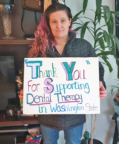 A woman stands in the living room of a house holding a sign that reads "Thank you for supporting dental therapy in Washington state."
