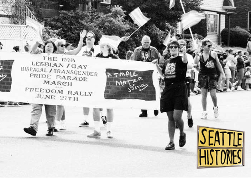 A black-and-white photo of people marching down a residential street carrying a banner reading "The 1993 Lesbian/Gay/Bisexual/Transgender Pride Parade March and Freedom Rally June 27th"