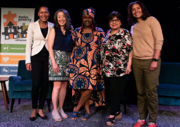 Five women of color stand together on a stage in front of two blue plush chairs. A Solid Ground banner with the hashtag #SolvePoverty can be seen behind them.