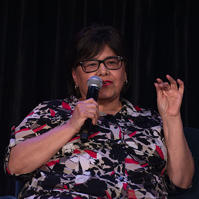 A women with dark hair and a red, black, and white patterned blouse gestures with one hand while holding a microphone in the other.