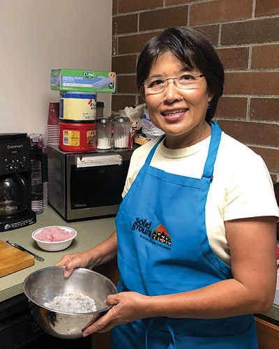 A smiling woman wearing glasses, a white T-shirt, and a blue apron holds a metal mixing bowl.