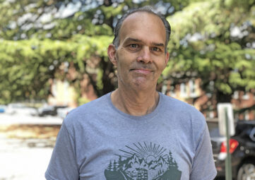 A man in a grey tshirt smiles at the camera. Trees can be seen out of focus in the background.