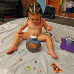 A toddler wearing only a diaper gazes down at a small pumpkin covered in blue painting, a paint brush held aloft in his hand.