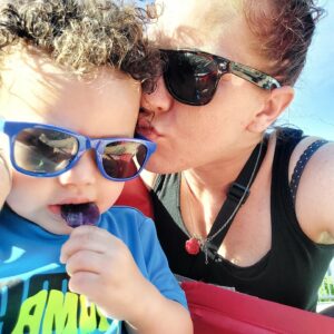 A woman in sunglasses kisses her young son on the side of the forehead.