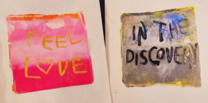 Two small paintings, one with a background painted in shades of pink, orange, and gold, and the other with a background of blue, black, and gold. The first has the words "feel love" painted in gold, and the second has the words "in the discovery" painted in black.