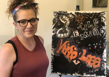 Woman smiles into the camera next to an easel with a multi-media art piece that reads: Exiled Mind/ Vivid Loss. She wears glasses, a red tank top, and her curly hair is pulled back with a headband.