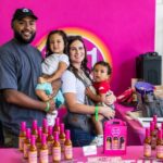 A man and woman holding two kids amid a display of hot sauces with bright pink labels and branding.