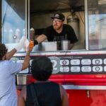 A man in a red food truck hands an order to a customer through a window.