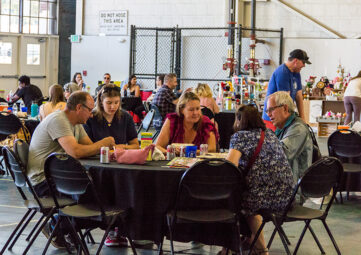 Five people talk and eat while seated at a table with a black table cloth in what looks in an airplane hangar. Other people can be see at similar tables in background.