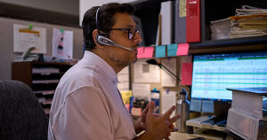 A man with dark hair, facial hair, and glasses wears a phone headset and faces a computer monitor as he talks on the phone.