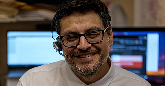 A man with dark hair, facial hair, and glasses smiles. He wears a phone headset and has computer monitors behind him.
