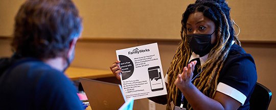 A white man and a Black woman sit across a table from each other, wearing masks. The woman has a laptop and holds up and explains a flyer about free cellphones.