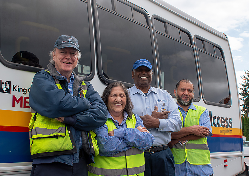 Four paratransit drivers, three men and one woman, stand smiling with arms crossed in from of a King County Metro ACCESS bus.