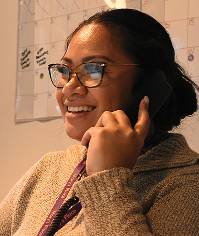 Young woman with dark hair pulled back smiles while talking on the phone. She wears glasses, a tan pullover and a purple Mary's Place lanyard.