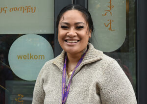 Young woman with dark hair pulled back smiles into the camera. She wears a tan pullover and a purple Mary's Place lanyard.