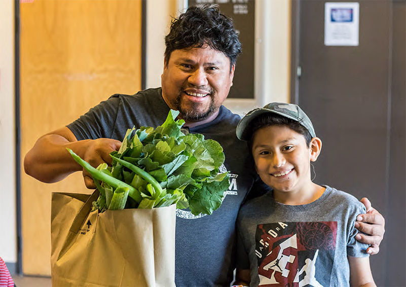 A smiling man holds up a brown paper grocery bag filled with vegetables with one hand and puts his other arm around his sons shoulder.