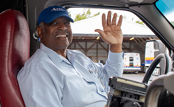 A Black man wearing a blue Access Transportation cap smiles and waves from the driver's seat of a Metro Access bus.