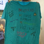 A person holds up a teal-colored shirt in front of them decorated with several phrases in Spanish, including "Amate" and "Alza tu voz."