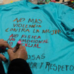 A close up of hands decorating a teal-colored shirt with the Spanish phrases, "No mass violencia contra law mujer. No fisica. No emocional. No sexual."
