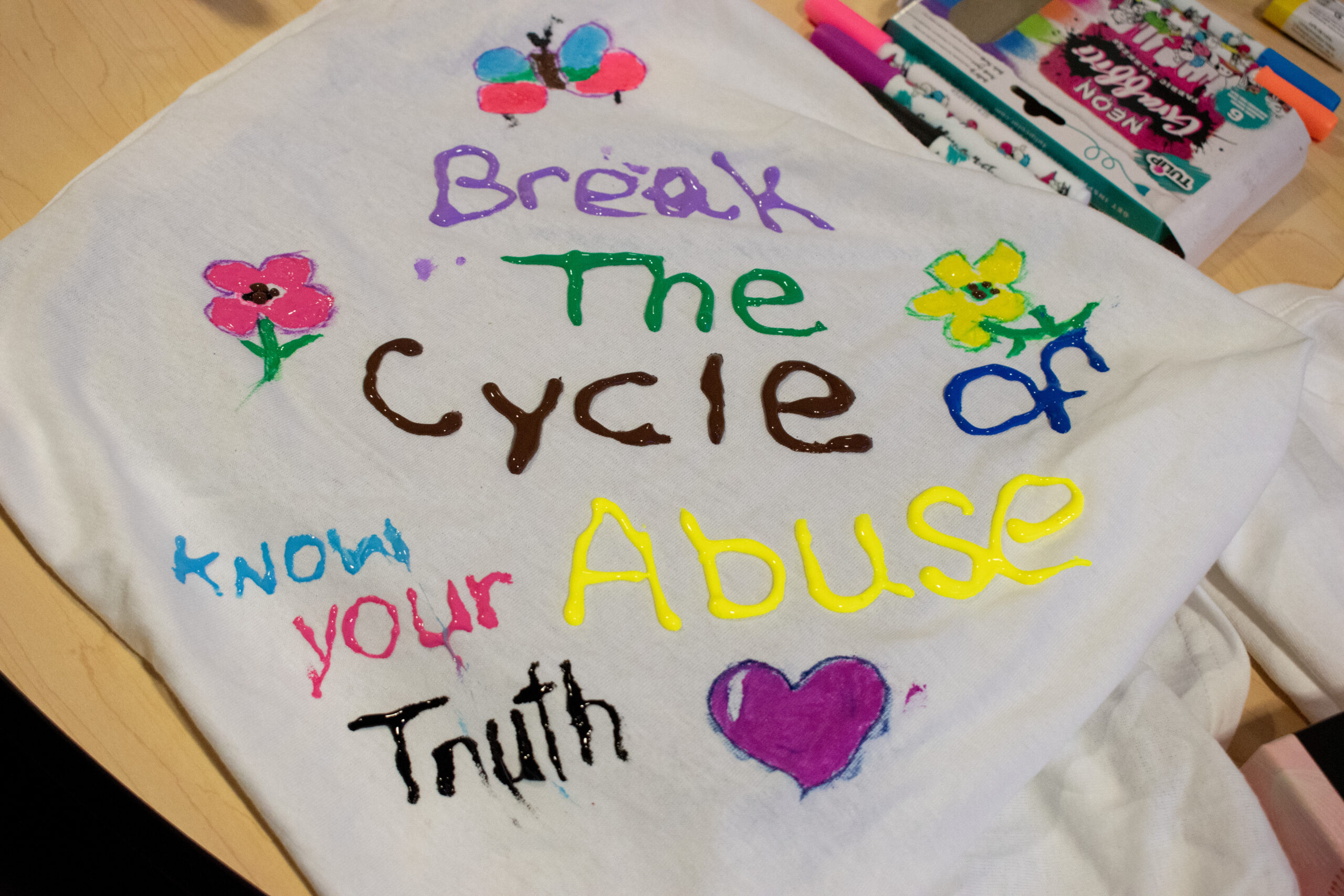 A white shirt painted with pictures of a heart, butterfly, and flowers, along with the phrases, "Break the cycle of abuse," and "Know your truth."