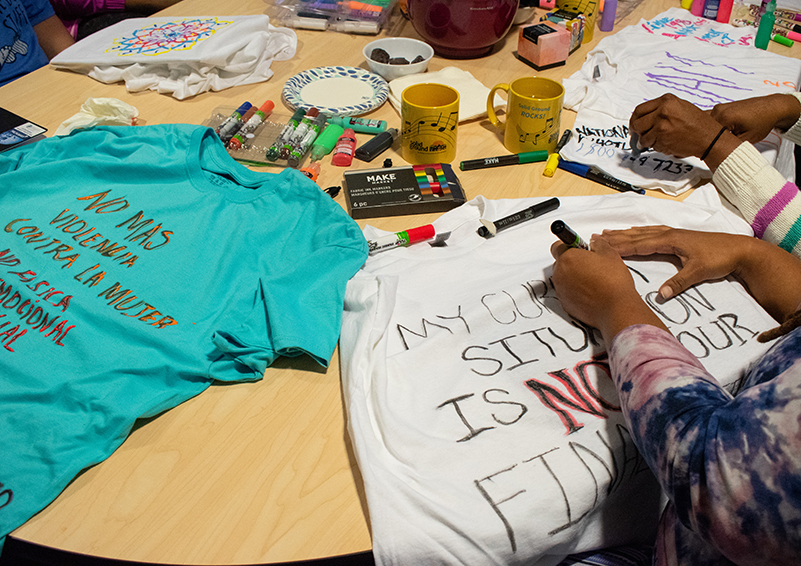 A close up shot of hands writing on and decorating shirts spread out on a table. A teal-colored shirt reads in Spanish, "No mas violencia contra la mujer."