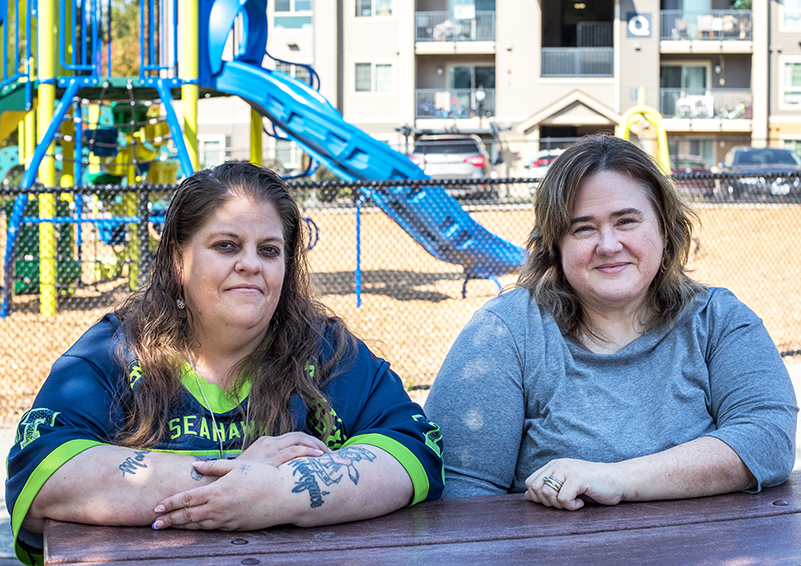 Two women, one in Seahawks jersey and the other in a gray long-sleeve shirt, sit at elbow to elbow at a picnic table with a playground and apartment building in the background.