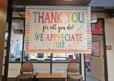 A colorful banner reading "Thank you for all you do! We Appreciate you!" hangs in an office.