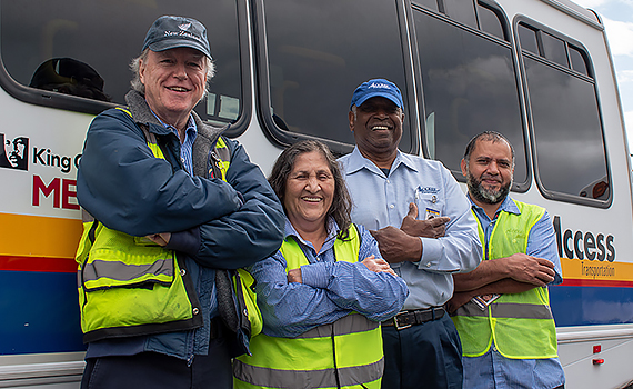 Four paratransit drivers, three men and one woman, stand smiling with arms crossed in from of a King County Metro ACCESS Transportation bus.